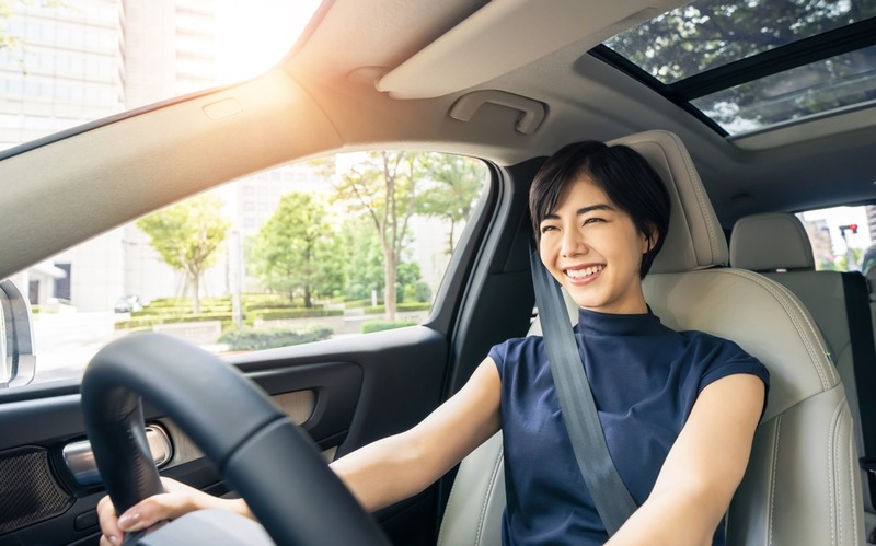 An attractive short dark-haired woman is sitting in the driver's seat of her GMC SUV smiling with her hands on the steering wheel, driving through a green neighborhood