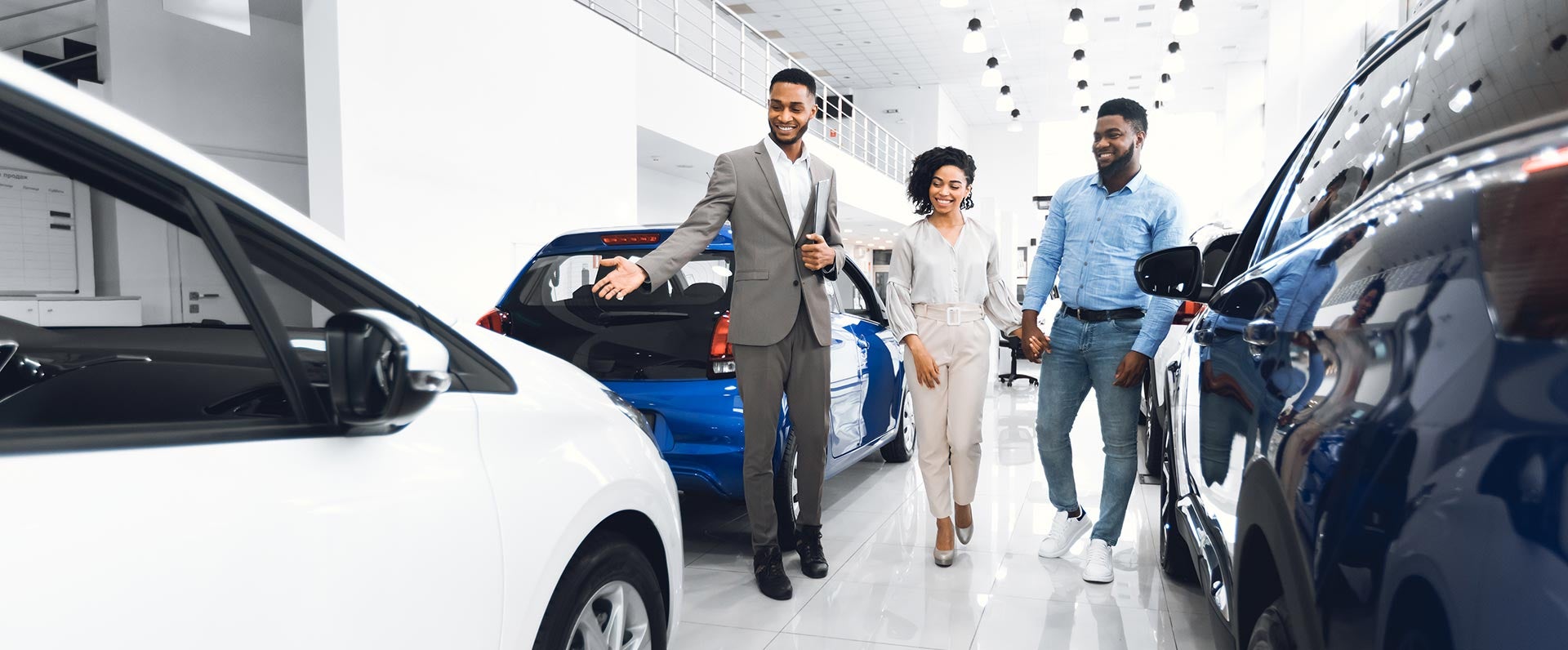 A suited GMC salesman is showing a couple his assortment of vehicles on the bright white showroom floor and they're all smiling, having a great time.